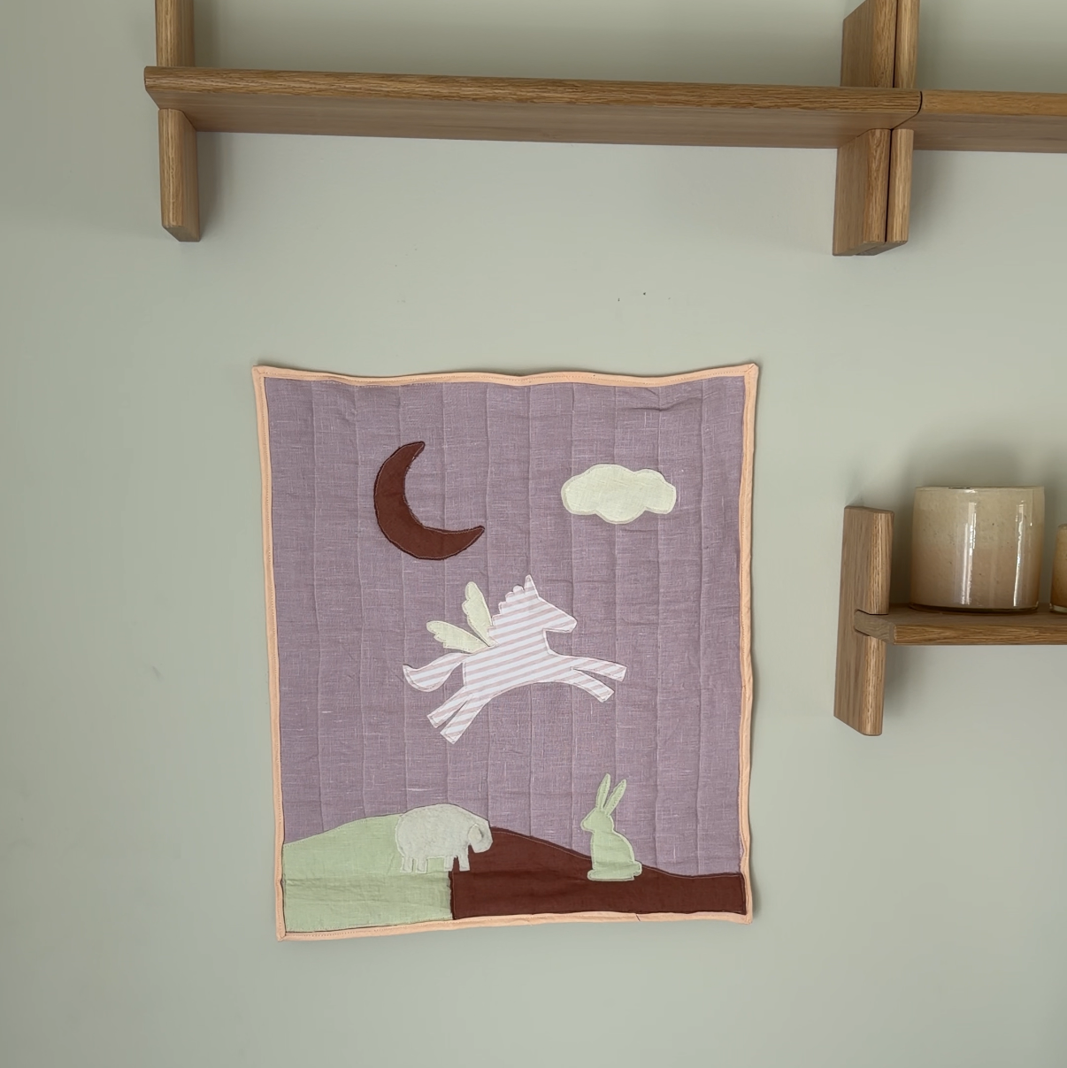 A small quilted wall hanging with a whimsical unicorn, moon, and cloud design in muted purple, cream, brown, and green tones, displayed on a light gray wall between two wooden shelves.