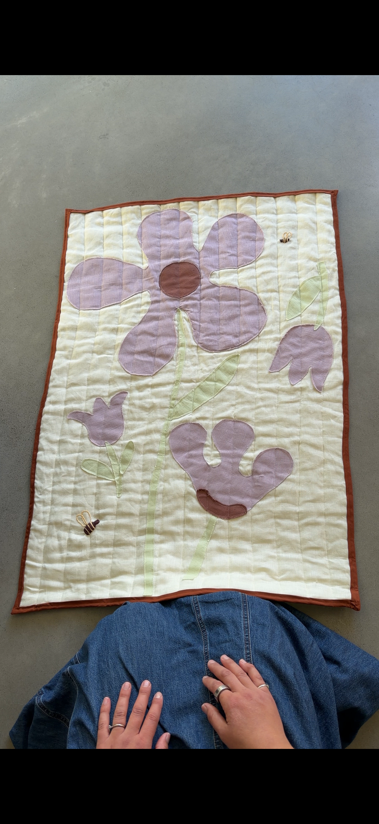A handmade quilted textile featuring purple flowers and green stems on a cream background, laid flat on a gray concrete floor with a person's hands resting on a blue denim garment below it.