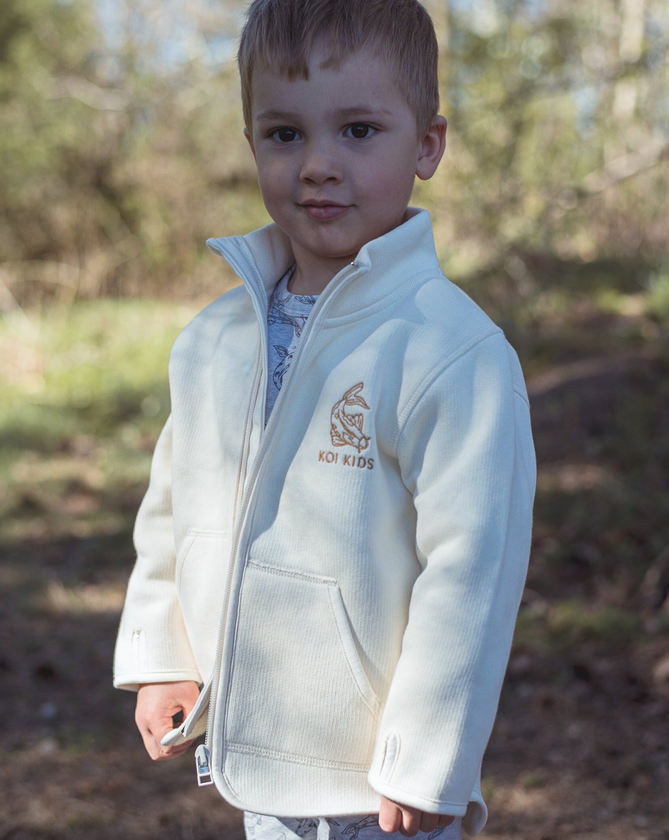 Offwhite jacket with zipper opening and pockets at front. Brown Koi fish logo embroidery at chest and sleeves with thumb holes.