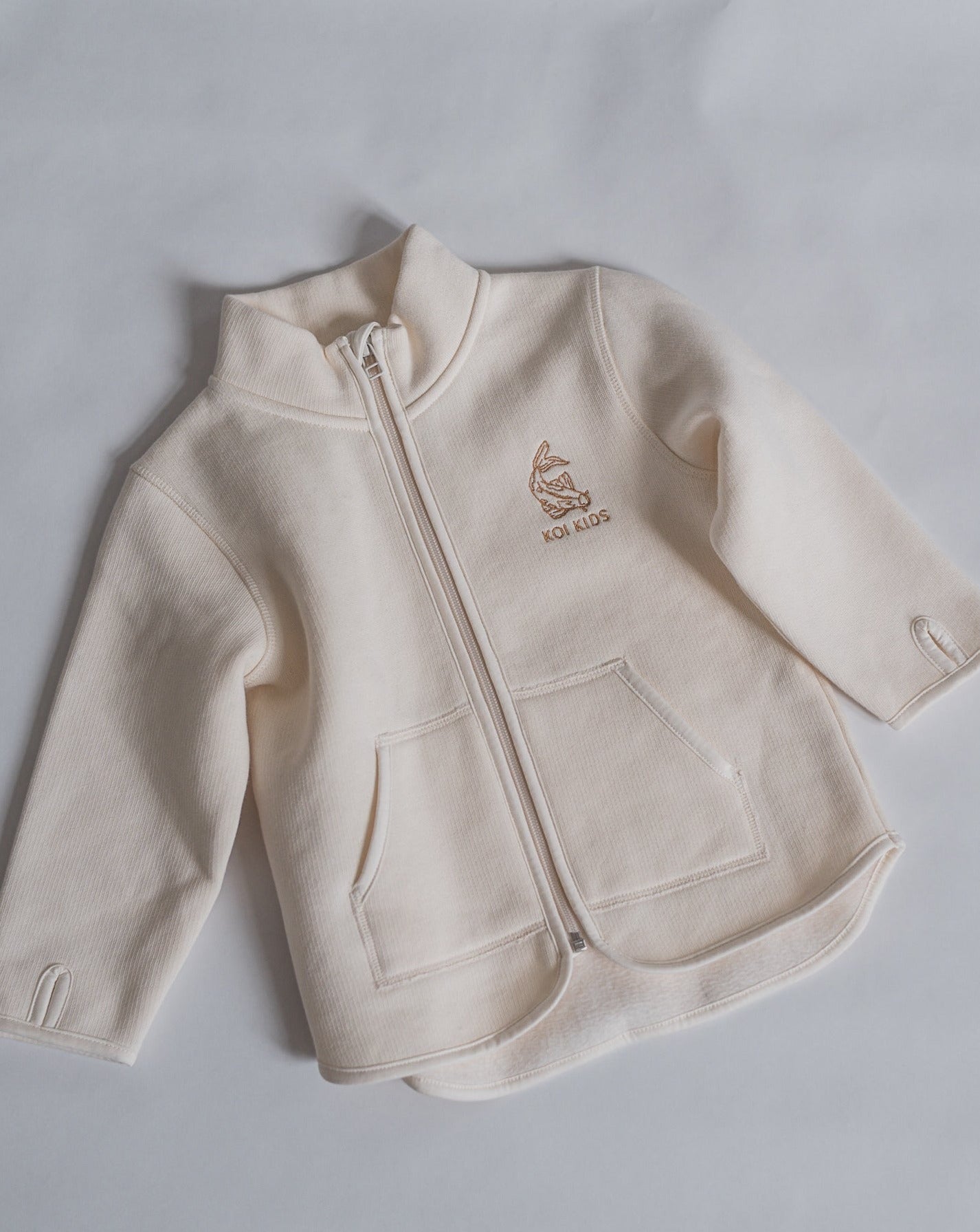 Offwhite jacket with zipper opening and pockets at front. Brown Koi fish logo embroidery at chest and sleeves with thumb holes.